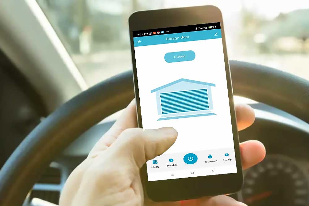 Hand holding a smartphone in a car, using a mobile app to check and control the status of a garage door remotely.